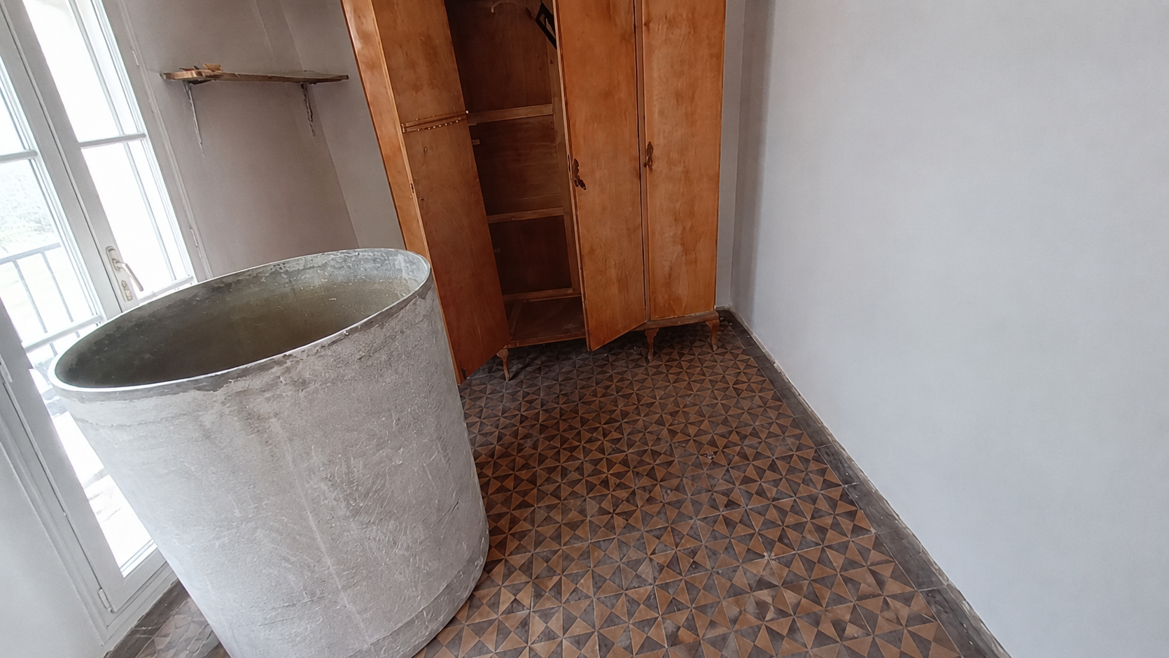 Habitación alargada con ventanales, bidón y suelo de baldosa antes de reformar